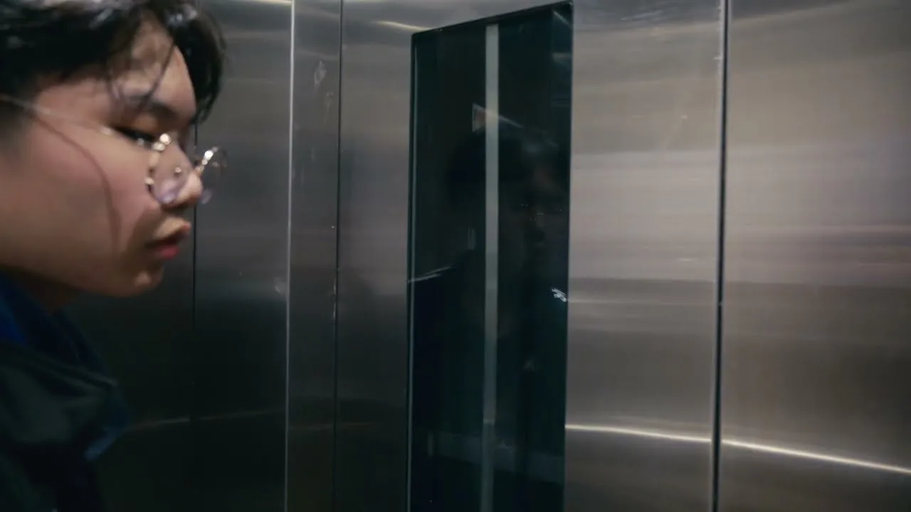 A boy in glasses stares at his reflection in steel lift doors